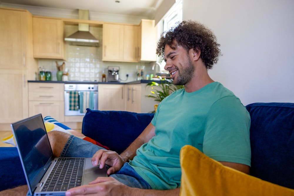 A side-view profile of a customer sitting on a sofa, with a laptop resting on their lap as they look down and smile at the screen.