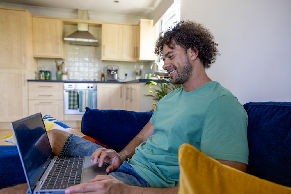 A side-view profile of a customer sitting on a sofa, with a laptop resting on their lap as they look down and smile at the screen.