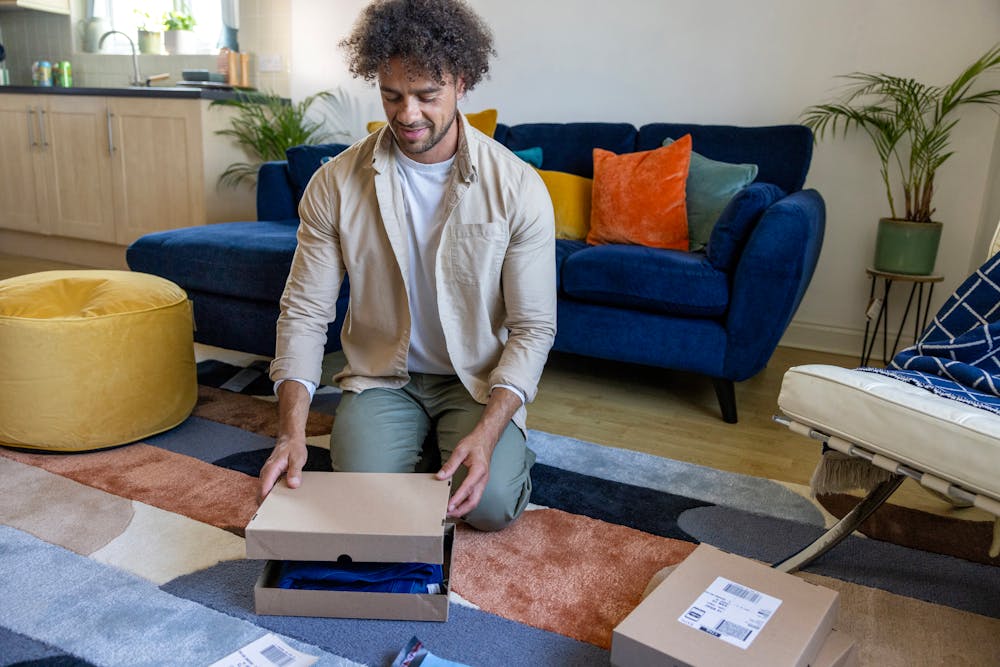 A customer is crouched on the floor of their apartment, holding items the lid of a box, ready to seal their parcel in front of them.