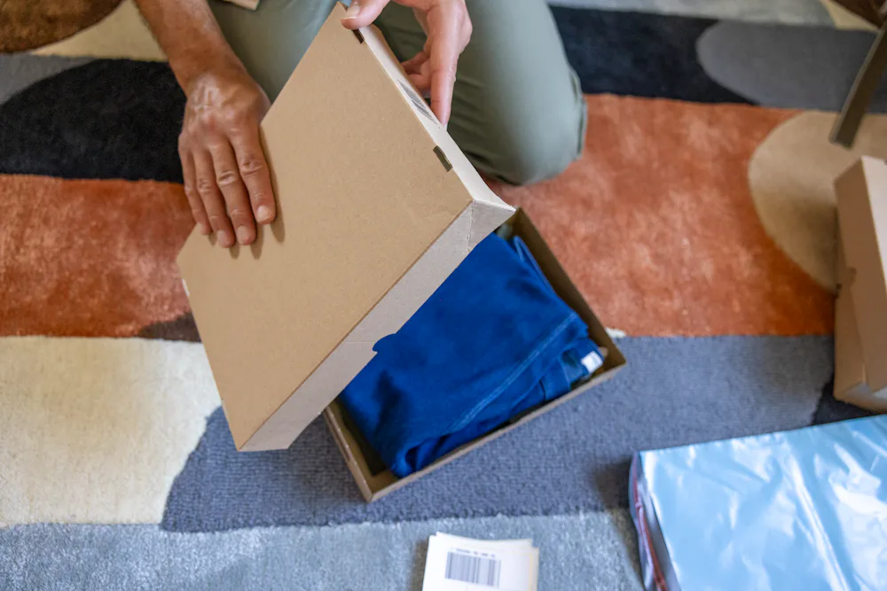A close-up of a customer's hands as they are crouched on the floor of their apartment, holding items the lid of a box, ready to seal their parcel in front of them.