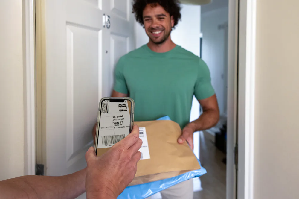 A customer is standing in the doorway of their home, holding their parcels and smiling. The courier's hand can be seen holding a device and scanning the parcel's barcode.