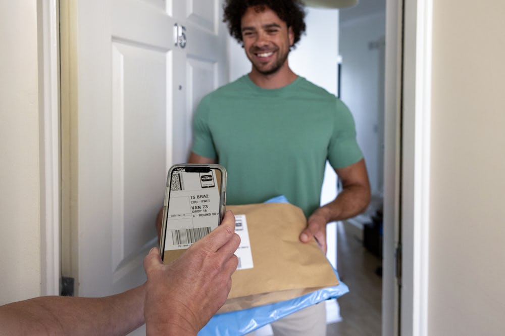 A customer is standing in the doorway of their home, holding their parcels and smiling. The courier's hand can be seen holding a device and scanning the parcel's barcode.