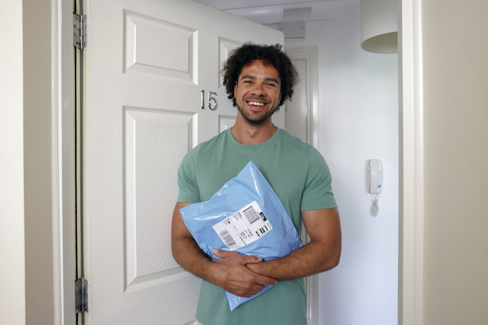 A customer is standing in the doorway of their home, holding their parcels and smiling for the camera.