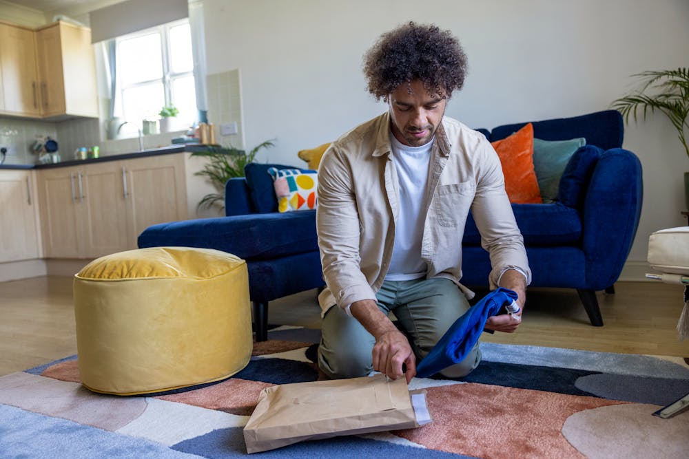A customer is crouched on the floor of their apartment, holding recyclable packaging in one hand and items in another.