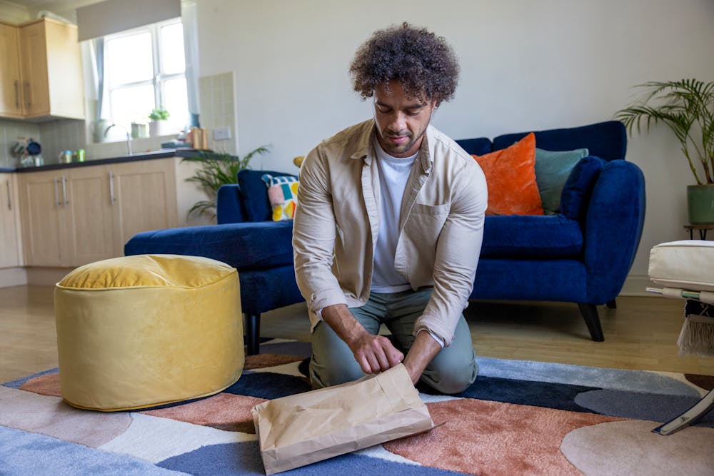 A customer is crouched on the floor of their apartment, holding recyclable packaging in one hand and inserting items with the other.