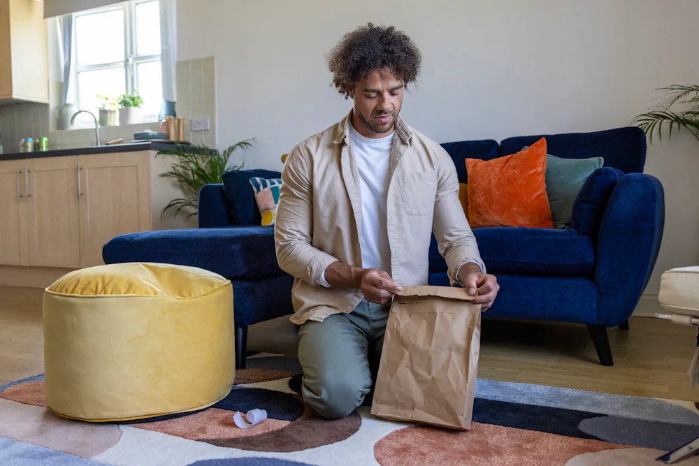 A customer is crouched on the floor of their apartment, sticking down the seal of their recyclable packaging.