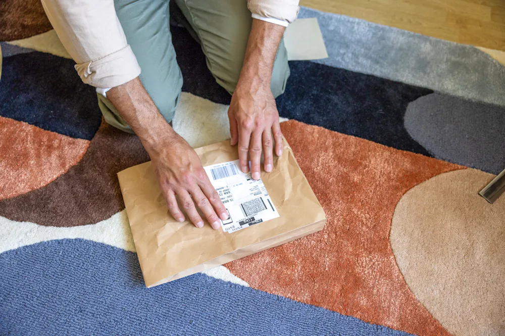A close-up of a customer's hands placing their label on the parcel in front of them and crouching down on the floor.