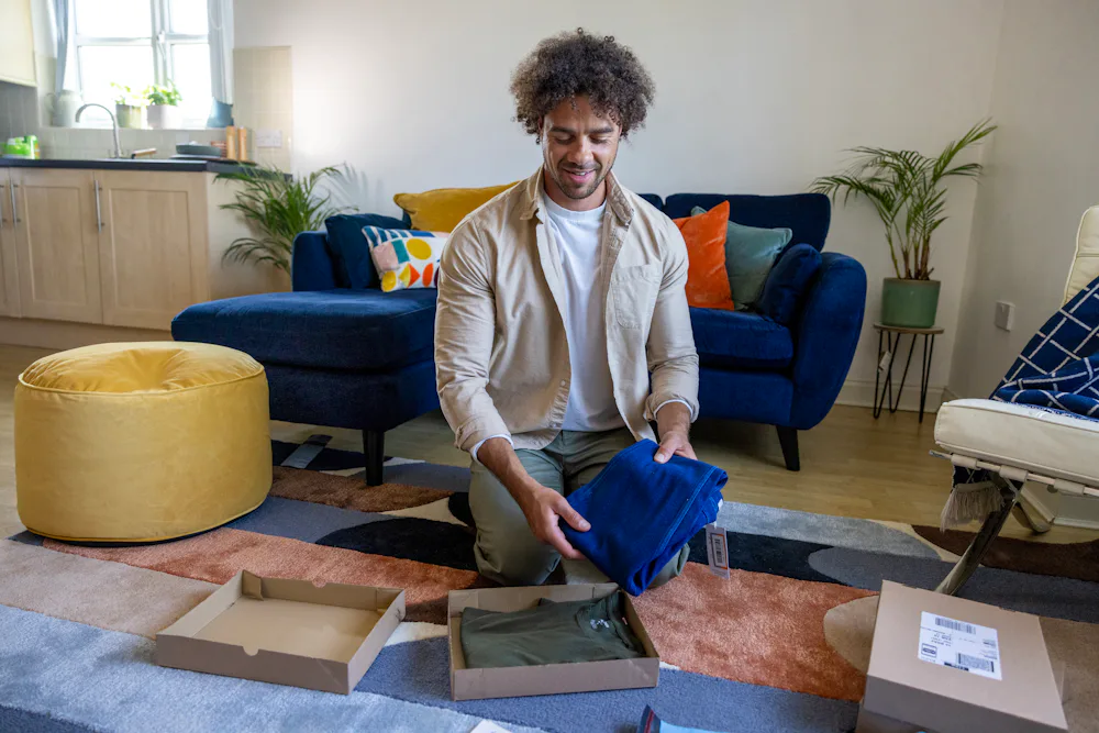 A customer is crouched on the floor of their apartment, holding items in one hand and is about to place them in the box in front of them.