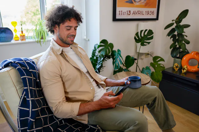 A customer sat on a sofa in their lounge, holding their mobile phone and looking at their screen with a mug in their other hand.