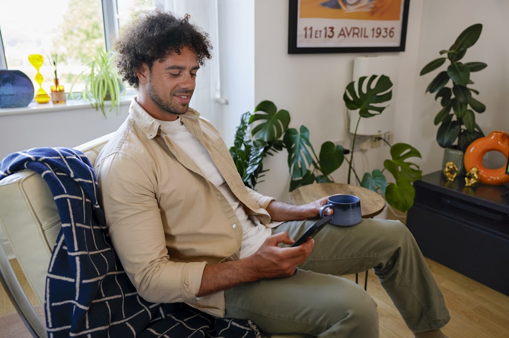 A customer sat on a sofa in their lounge, holding their mobile phone and looking at their screen with a mug in their other hand.