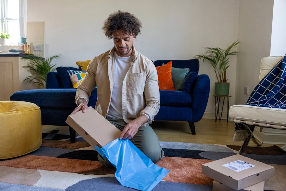 A customer is crouched on the floor of their apartment, placing a cardboard parcel inside waterproof packaging.