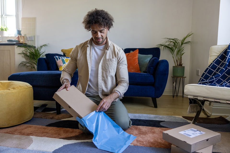 A customer is crouched on the floor of their apartment, placing a cardboard parcel inside waterproof packaging.