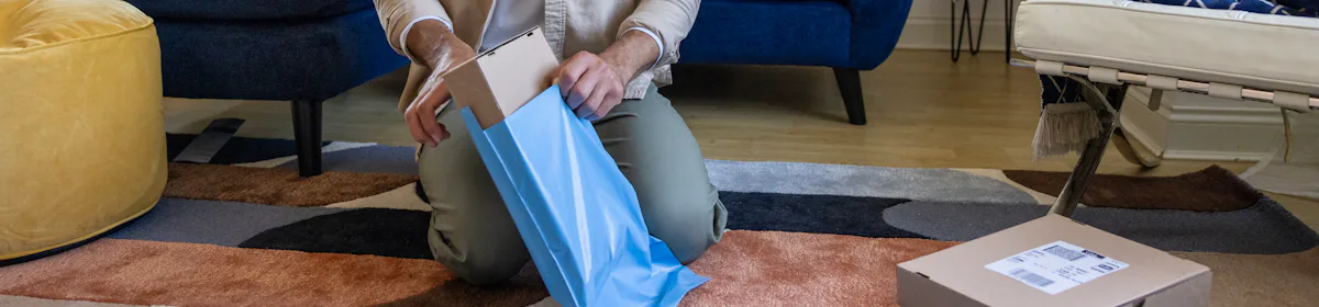 A customer is crouched on the floor of their apartment, placing a cardboard parcel inside waterproof packaging.