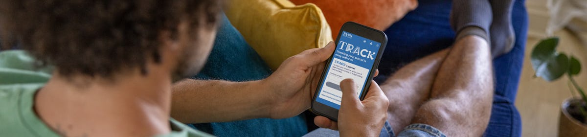 A close-up of a customer sitting on a sofa, holding their phone and looking at the screen. The display shows the 'Track' page for the customer to track where their parcel is on its journey.