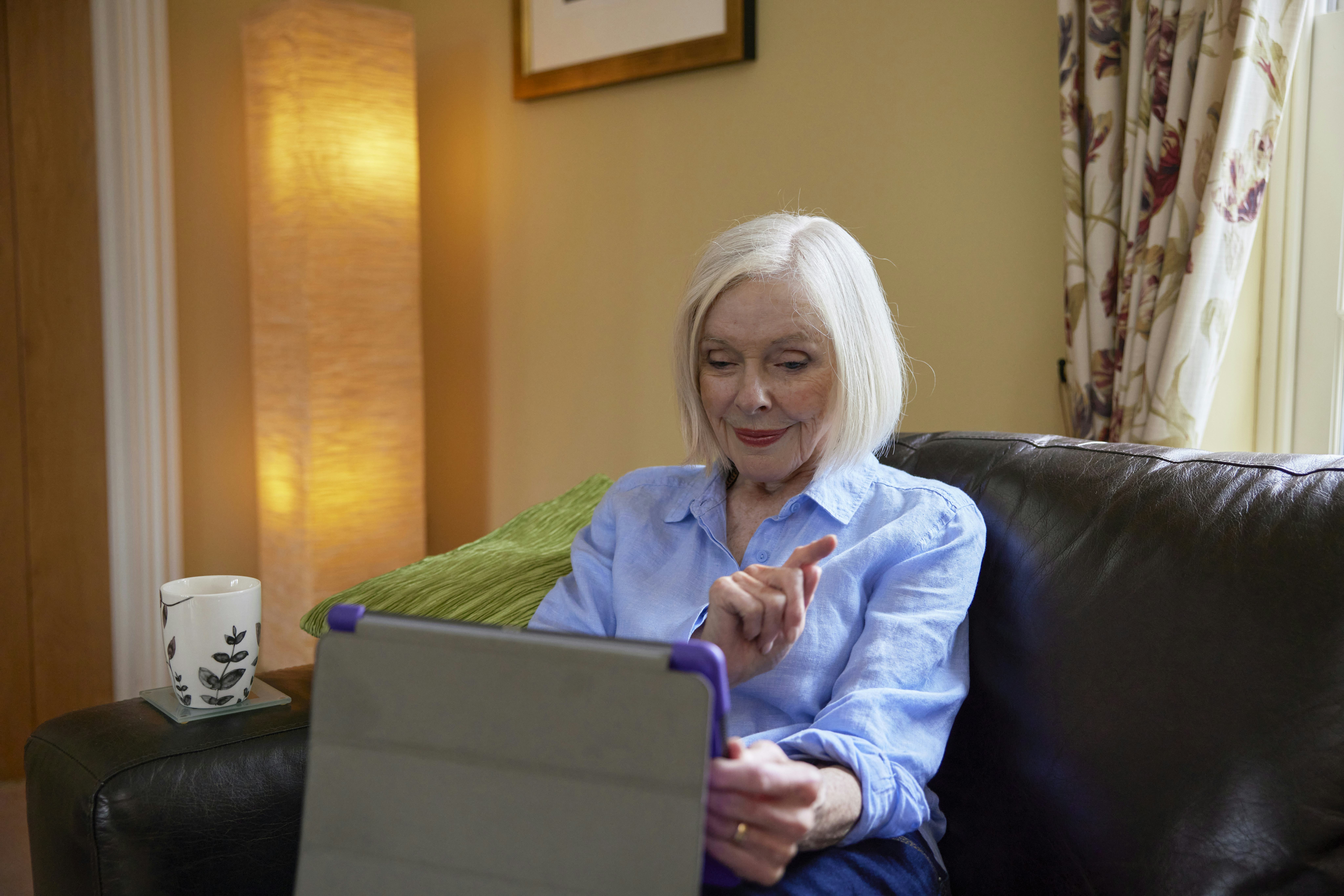 An older woman sits on a brown sofa. A cup of tea is resting of the arm. She is using an Ipad tablet to order a parcel on the Evri website.