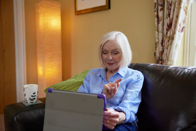 An older woman sits on a brown sofa. A cup of tea is resting of the arm. She is using an Ipad tablet to order a parcel on the Evri website.