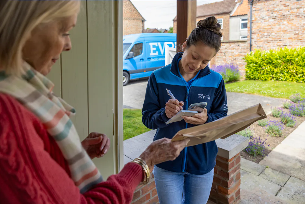 An older woman hands a parcel to a courier for collection. The courier is signing a blue slip. Blue Evri van visible in the background.
