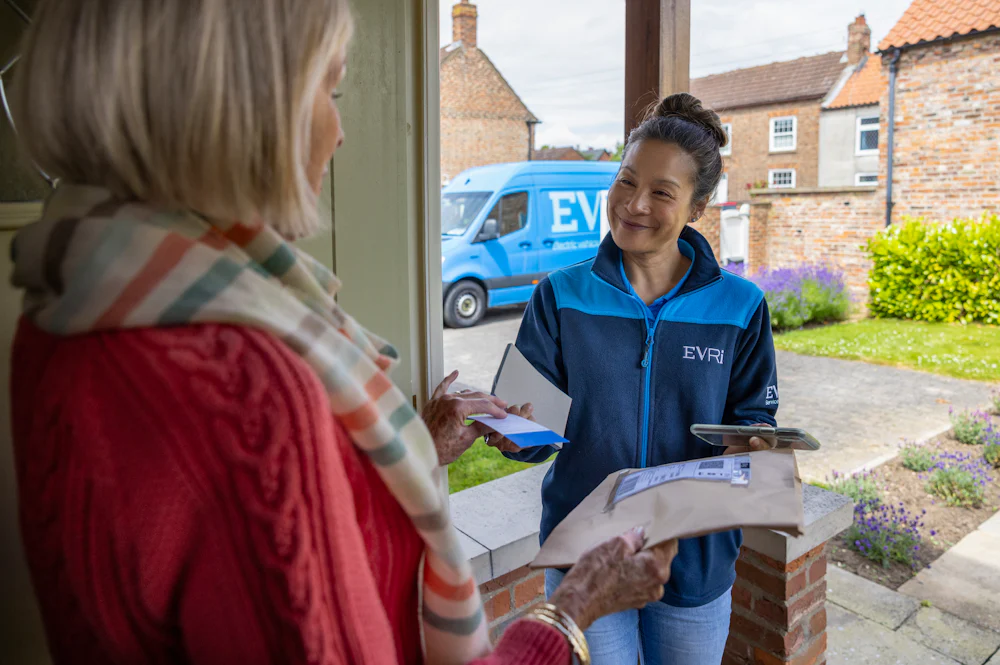 A courier is handing a blue collection receipt to an older woman at her door. Blue Evri van visible in the background.