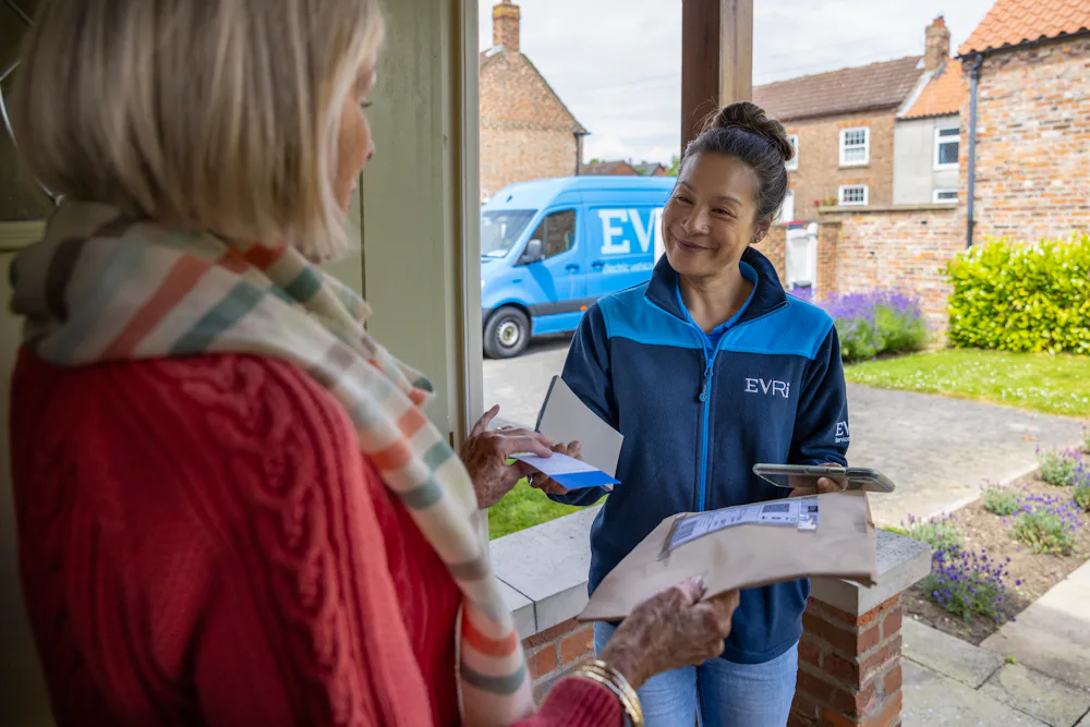 A courier is handing a blue collection receipt to an older woman at her door. Blue Evri van visible in the background.