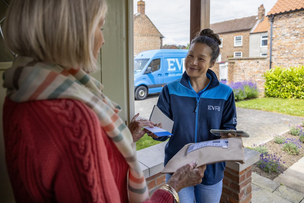 A courier is handing a blue collection receipt to an older woman at her door. Blue Evri van visible in the background.