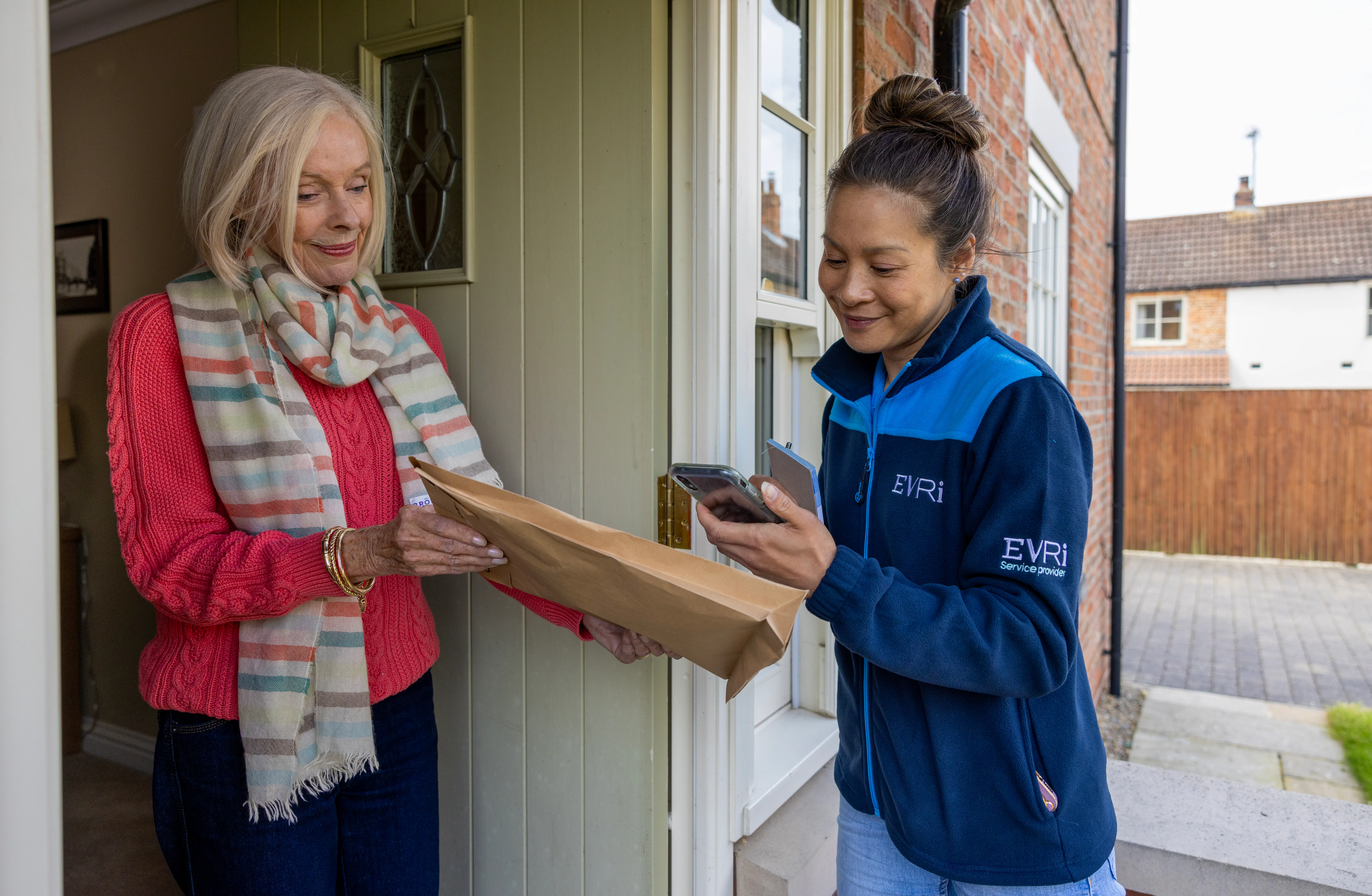 An older woman hands a parcel to a courier for collection. The courier is taking a photo.
