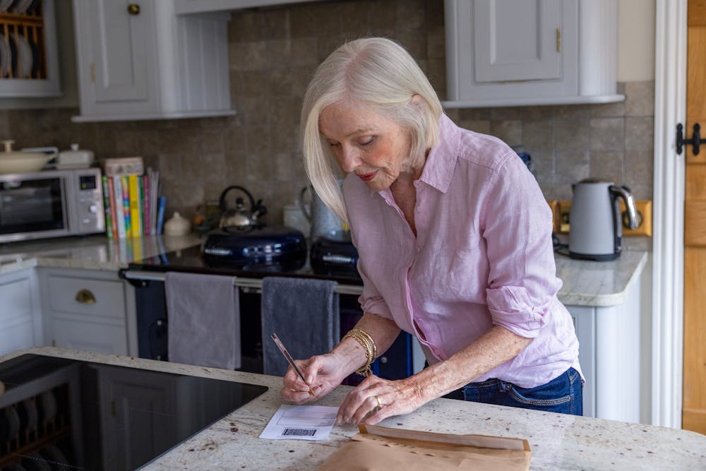 An older woman is writing on a label. A wrapped parcel is visible on the table in front of her.