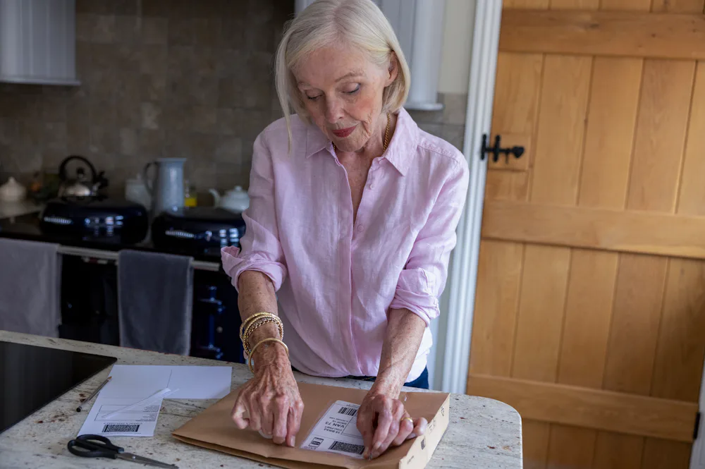 An older woman is wrapping a parcel. She is applying the label to her parcel with tape.