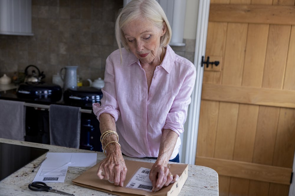 An older woman is wrapping a parcel. She is applying the label to her parcel with tape.
