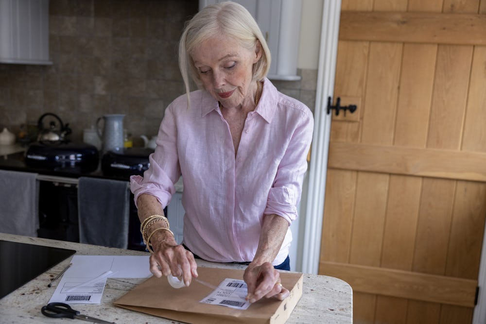 An older woman is wrapping a parcel. She is applying the label to her parcel with tape.