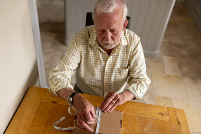An older man is sat a table. He is measuring a parcel's length with a tape measure.