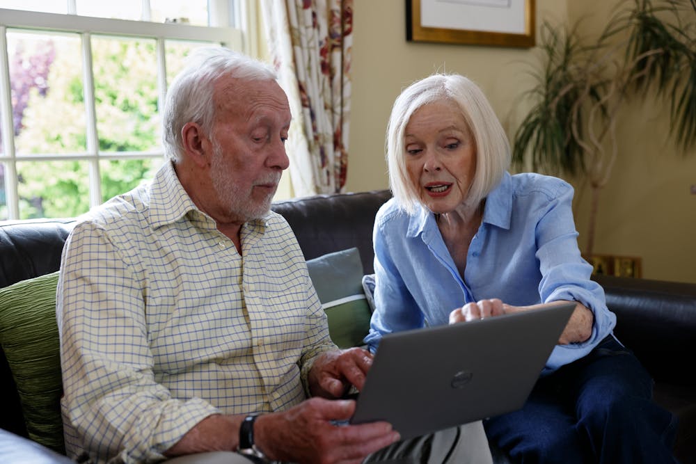 An older couple, a man and a woman, are sat on a sofa. The woman is showing the man something on the screen.