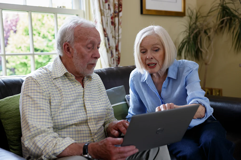 An older couple, a man and a woman, are sat on a sofa. The woman is showing the man something on the screen.
