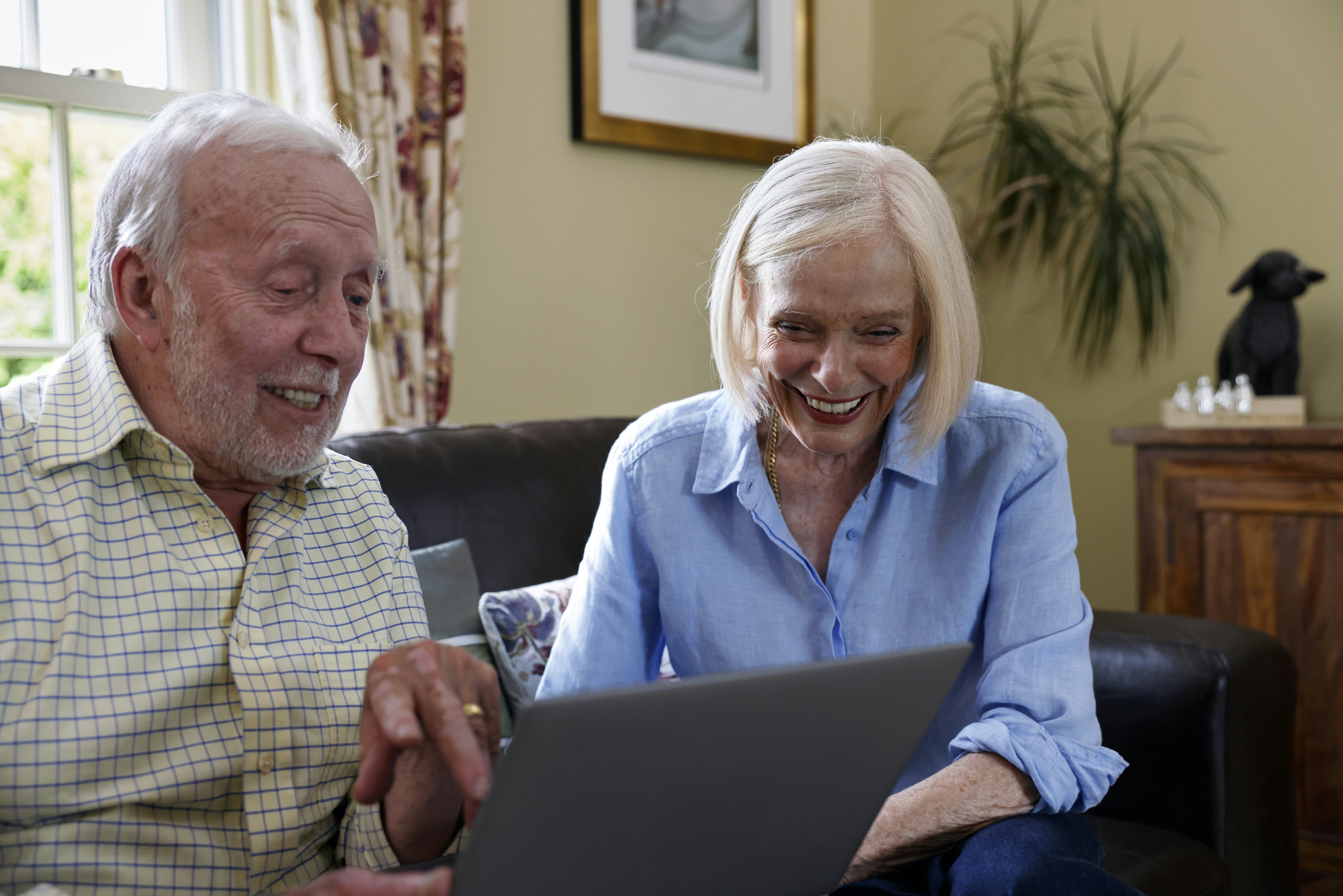 An older couple, a man and a woman, are sat on a sofa. They are laughing whilst using a laptop.