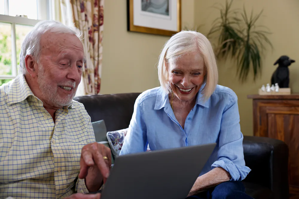An older couple, a man and a woman, are sat on a sofa. They are laughing whilst using a laptop.