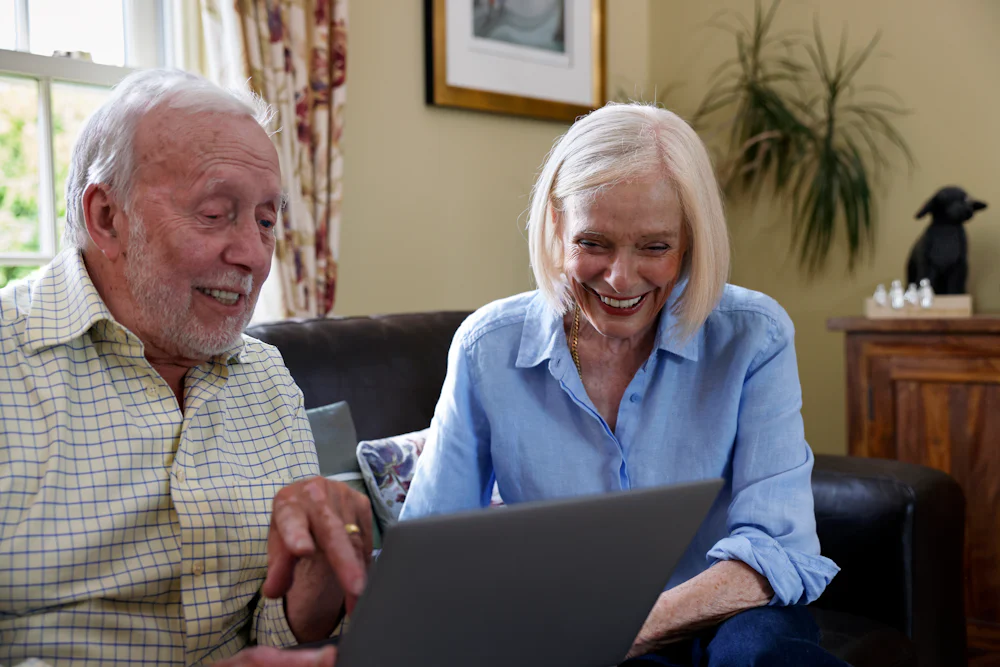 An older couple, a man and a woman, are sat on a sofa. They are laughing whilst using a laptop.