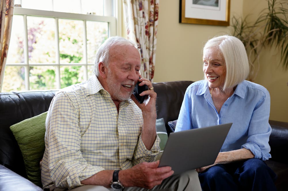 An older couple, a man and a woman, are sat on a sofa. The man is on the phone while the woman sits beside him. They look happy.