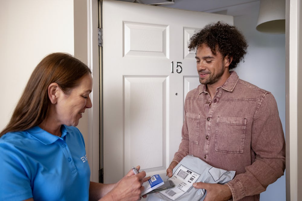A customer is handing an Evri Courier their parcel during a doorstep collection in a block of flats. The Courier is filling in the details of the collection on a blue slip to hand to the customer
