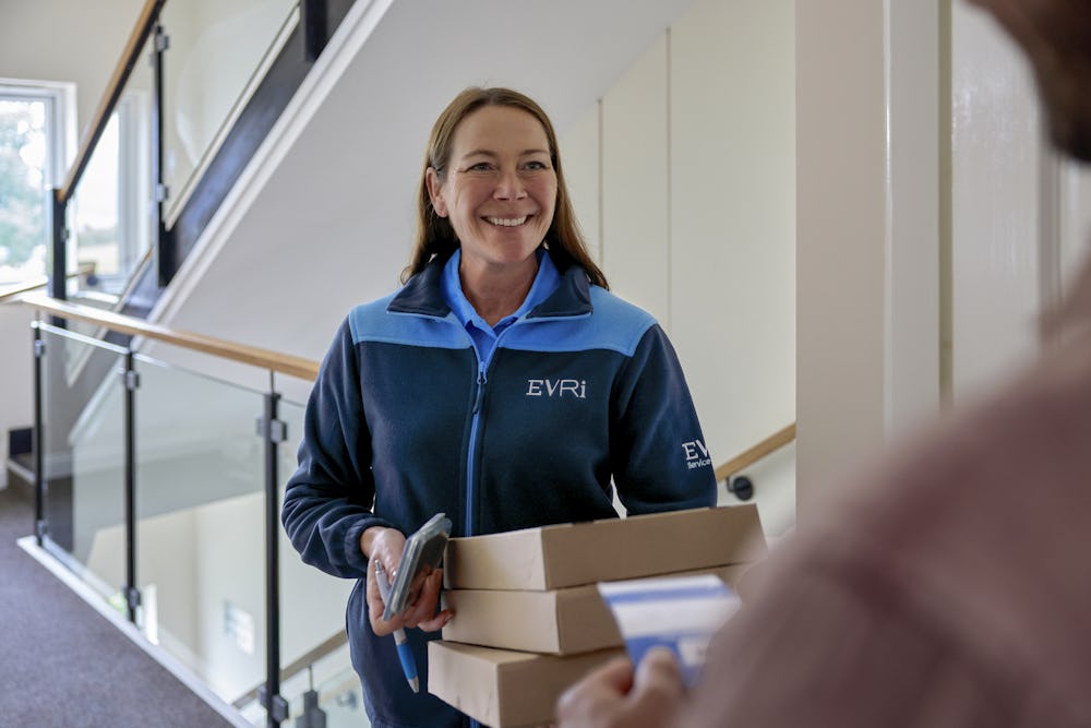 An Evri Courier smiling towards a customer after having completed a doorstep collection. The courier is holding multiple parcels, whilst the customer is holding a blue slip containing the details of the delivery.