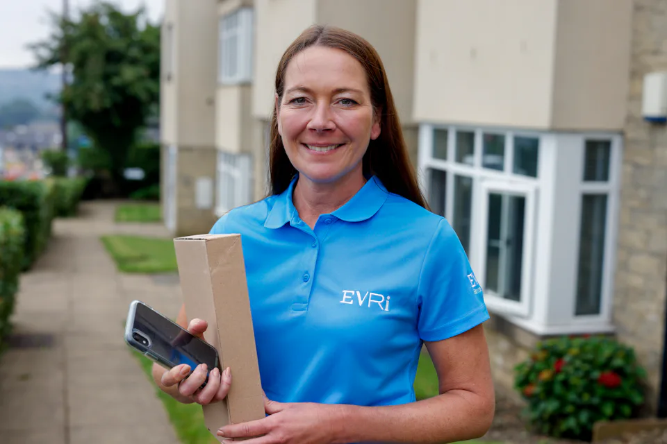 An Evri courier smiling and posing for the camera holding a parcel and their mobile device.
