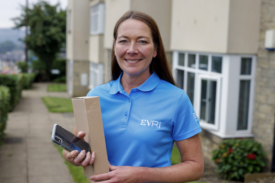 An Evri courier smiling and posing for the camera holding a parcel and their mobile device.