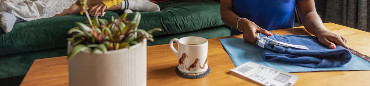 A mother is folding a pair of jeans on a coffee table, preparing them to be packaged and sent with Evri. Her two children are sat talking to eachother on the sofa behind her.