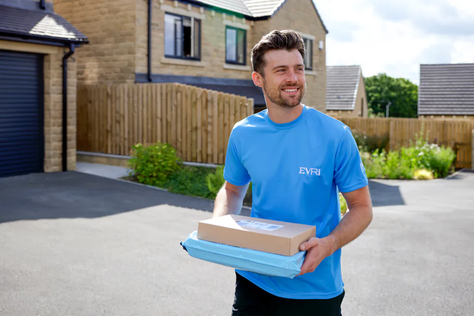 An Evri courier walking down a driveway in a suburban neighbourhood carrying two parcels.