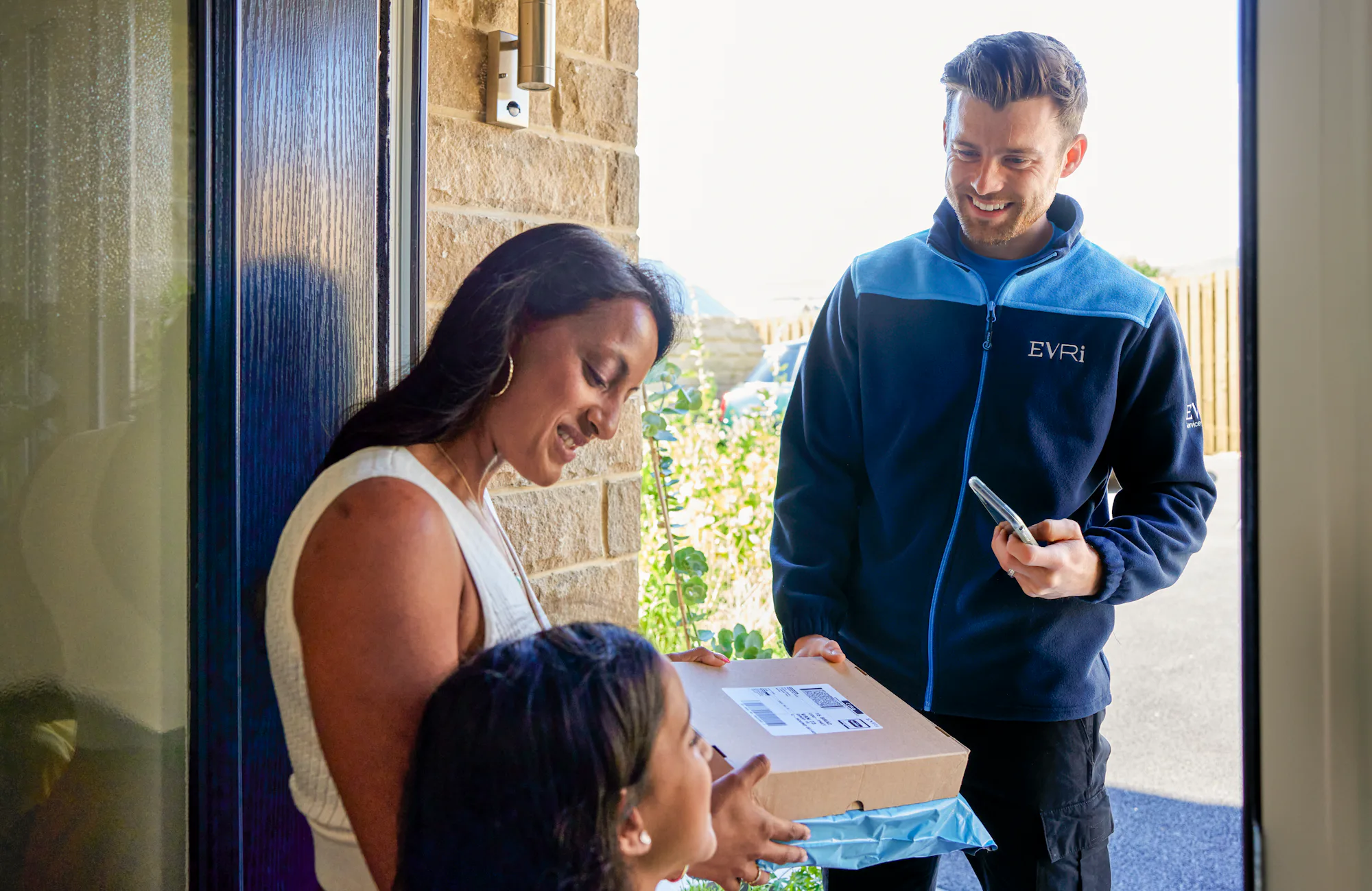 An Evri Courier handing over two parcels to an mother and child at their doorstep. The three are laughing during a pleasant exchange