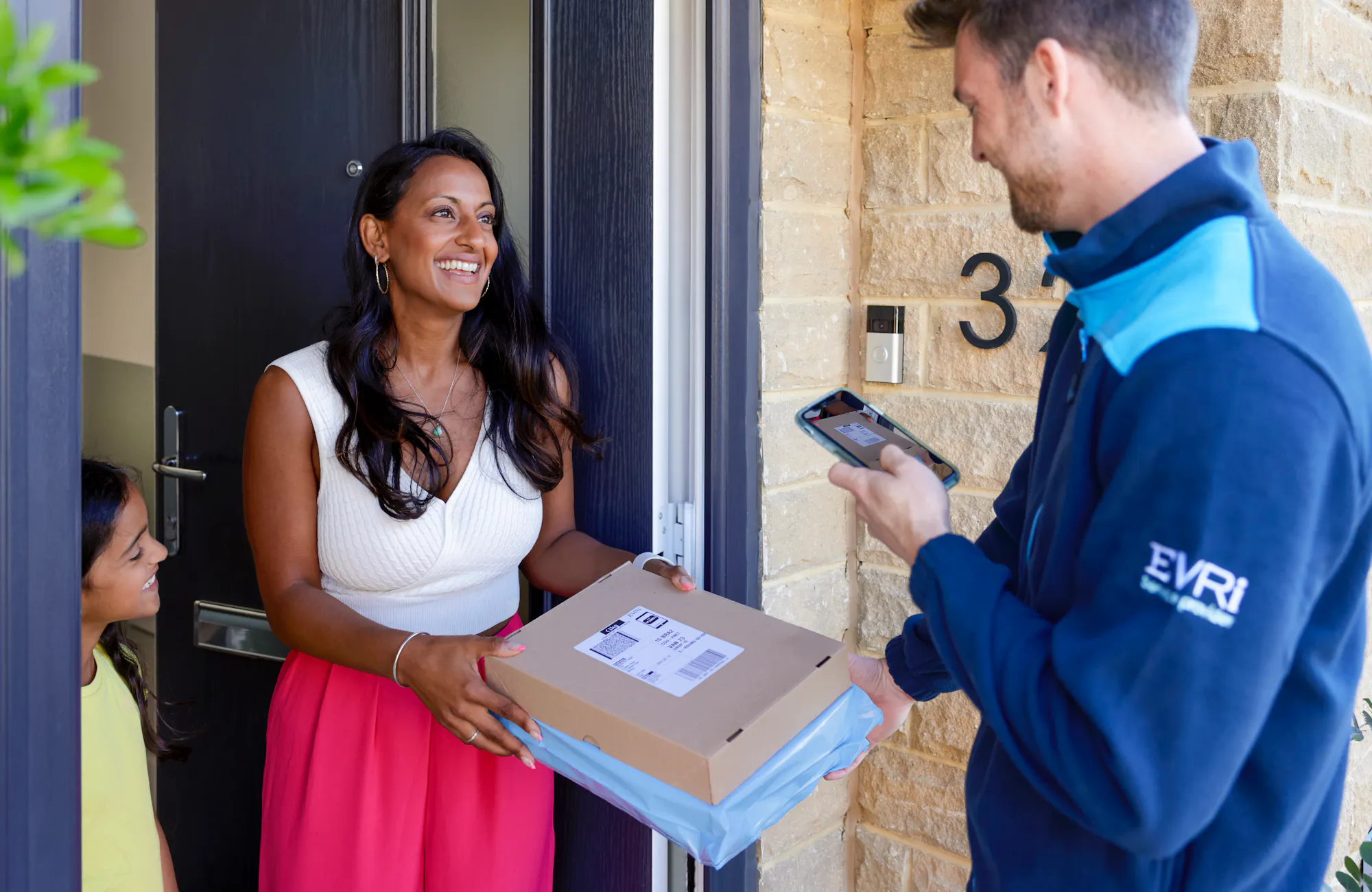 An Evri Courier delivering a parcel to an excited mother and child. The Courier is taking a photo for proof of delivery on their mobile device.