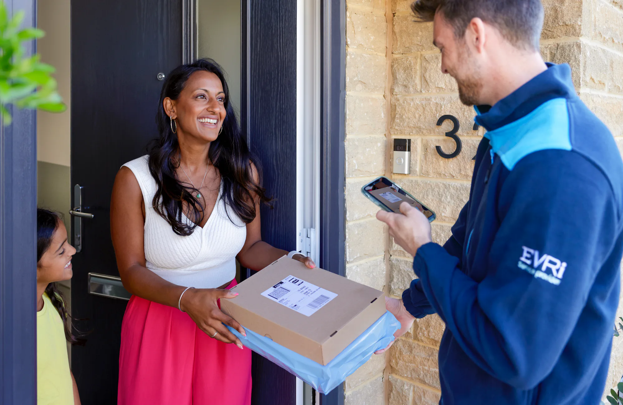 An Evri Courier delivering a parcel to an excited mother and child. The Courier is taking a photo for proof of delivery on their mobile device.