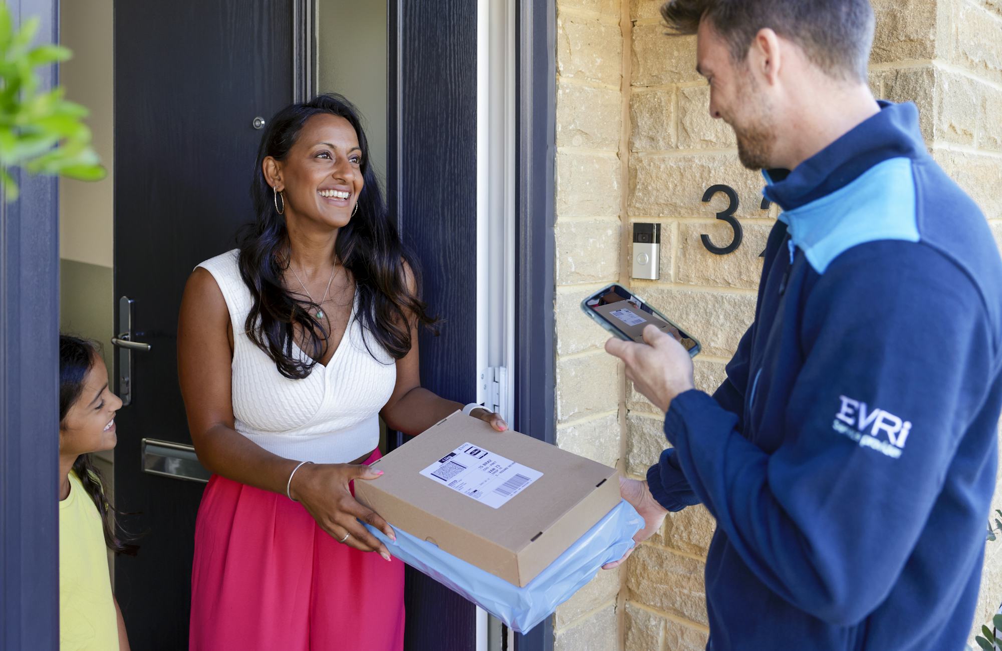 An Evri Courier delivering a parcel to an excited mother and child. The Courier is taking a photo for proof of delivery on their mobile device.