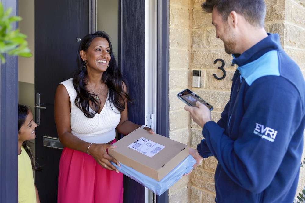 An Evri Courier delivering a parcel to an excited mother and child. The Courier is taking a photo for proof of delivery on their mobile device.