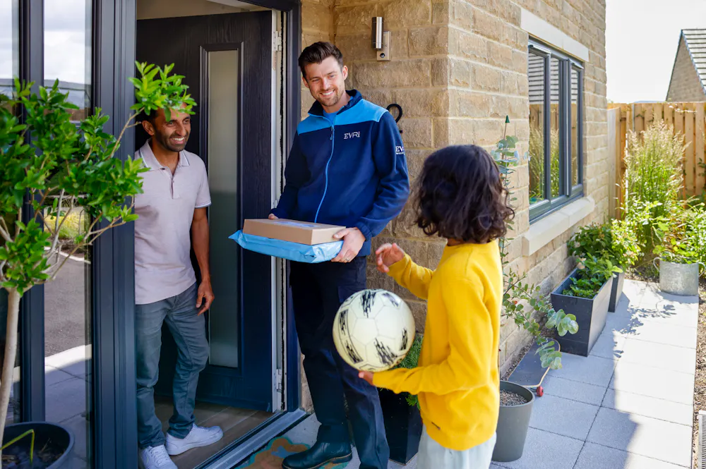 An Evri Courier is stood at a doorway holding two parcels. They are engaging in joyful conversation with a customer and a child with a football during the delivery.