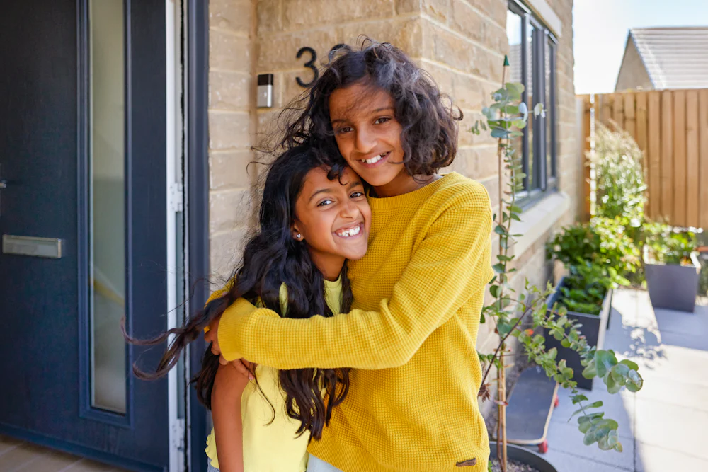 Two children are smiling and embracing eachother in front of their house whilst posing towards the camera.