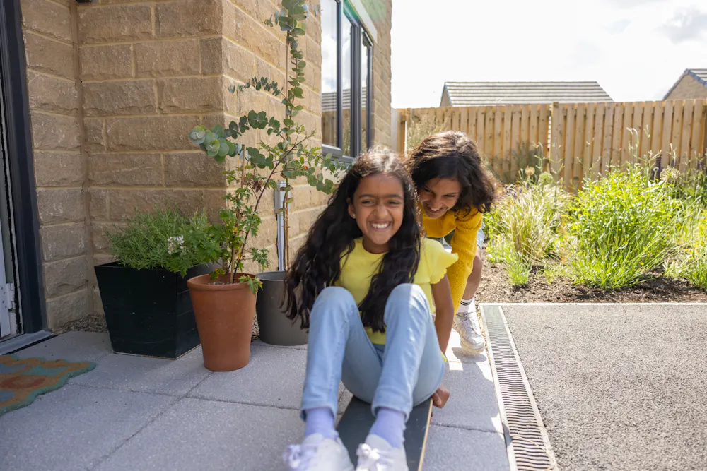 A child is pushing their sibling on a skateboard in front of their doorstep. They are both laughing and acting playful.